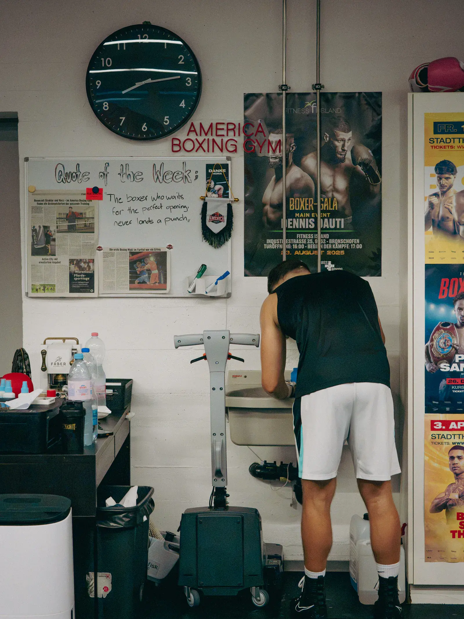 Wand im Gym mit grosser Uhr, dem Schriftzug America Boxing Gym, einer Pinnwand mit dem Zitat der Woche und Boxplakaten.