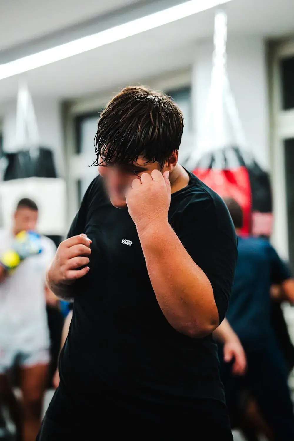 Junge im schwarzen T-Shirt mit erhobenen Fäusten im Kids-Training, dahinter Sandsäcke und weitere Kinder.
