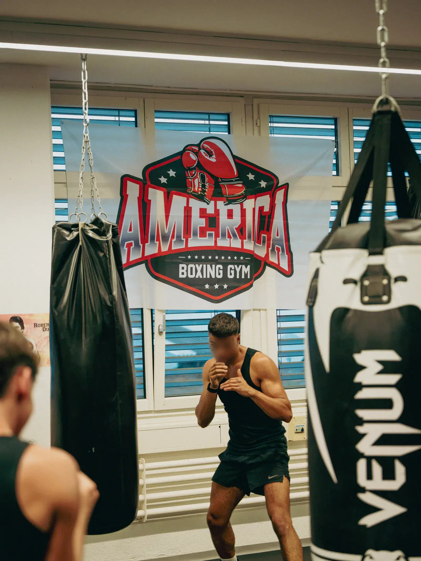 Boxer beim Schattenboxen zwischen zwei Sandsäcken, im Hintergrund das Banner des America Boxing Gym.
